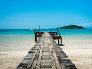 Scenic view of long rustic wooden pier bridge over crystal clear turquoise water and white sand beach of Koh Mak Island with Koh Kham Island at horizon, Trat, Thailand.