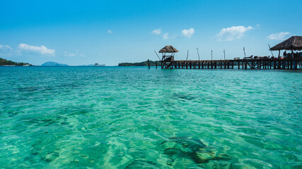 Koh Mak Island, Trat, Thailand. Scenic view of peaceful sea, crystal clear turquoise water with coral reef transparent and long wooden pier bridge landmark against blue sky in summer.