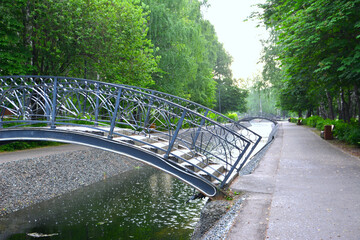 metal bridge across water tunnel in public park with green trees and paths in summer rainy day