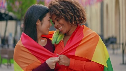 Multicultural lesbian couple wrapped in lgbt flag, tenderly hugging and smiling