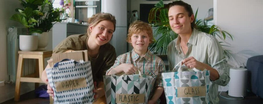 Portrait of happy lesbian mothers and their little son holding bags with paper, plastic and glass, looking at camera and smiling while sorting waste at home