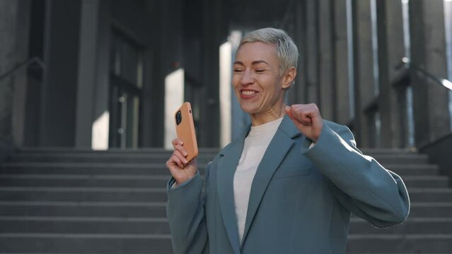 Joyful caucasian business woman with smartphone in hands emotionally gesturing while celebrating her success at work. Victory, lottery, technology and people concept.