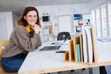 young business woman sits at a desk with laptop and laughs into the camera