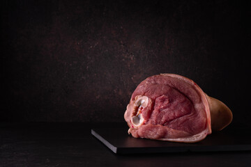 fresh raw pork knuckle lies whole on a black cutting board on a dark soft background. moody artistic food still life in a minimalist style