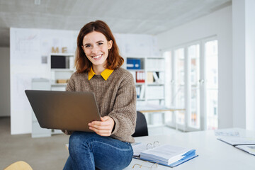 young smiling business woman sitting on a desk with laptop in her hands - 495080535