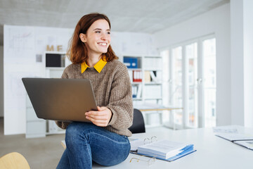 young happy business woman sitting on a desk with laptop in her hands - 495080564