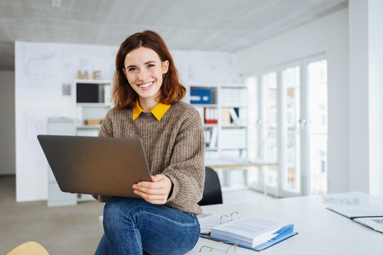 young smiling business woman sitting on a desk with laptop in her hands