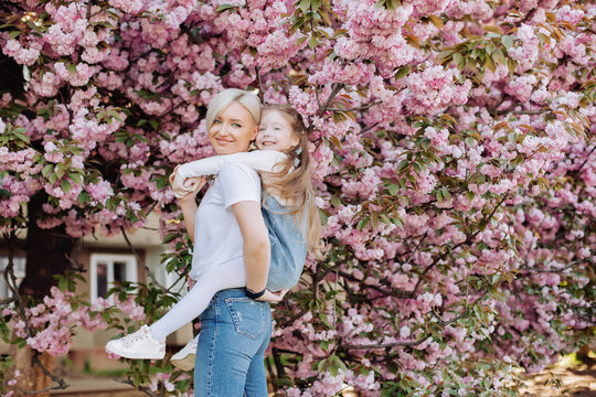 People, holidays and family concept - daughter hugs mom sitting on her back on cherry blossom background. Walk in the sakura trees in the city.