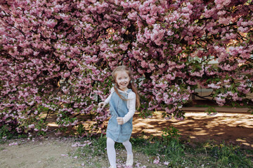 Beautiful red-haired girl sincerely smiles in the park near the sakura. Girl on a spring city street with blooming pink Japanese cherry trees.