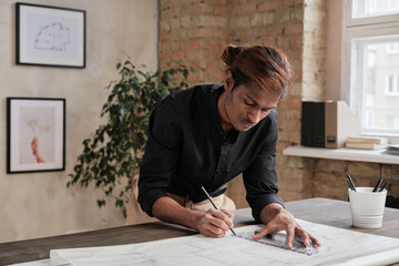 Serious young black architect with ponytail leaning on wooden table and drawing sketch using ruler in loft office