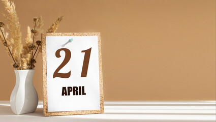 April 21. 21th day of month, calendar date.White vase with dead wood next to cork board with numbers. White-beige background with striped shadow. Concept of day of year, time planner, spring month