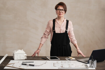 Portrait of content attractive mature lady in black bodysuit and eyeglasses standing at table with floor plan and D model of house
