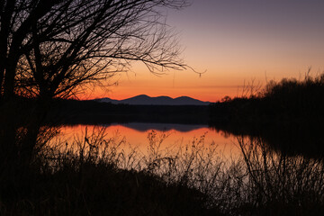 Landscape of mountain reflecting in river water framed with reeds and bare tree branches silhouettes at riverbank at dusk with clear sky and orange glow at horizon