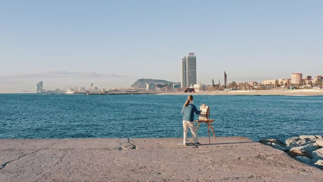 Focused lady artist freelancer paint with oil on canvas on plein air stand on stone slabs in front of splendid harbor of Barcelona city with modern skyscrapers above blue sea. 360 degree panorama view