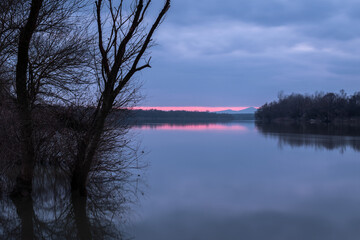 Landscape of river, reflection of distant mountain in calm water and silhouette of tree with bare branches at shore during overcast dusk with purple glow at horizon