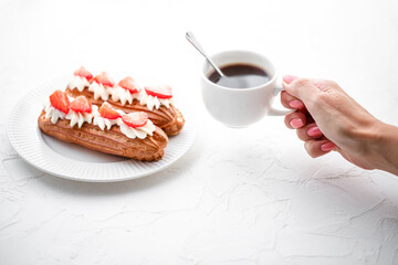 Woman's hand holding a cup of coffee over Eclairs with cream and strawberries on a white table