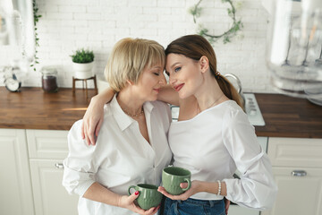Portrait of a happy elderly mother and daughter in the kitchen, they drink tea and enjoy a conversation. Tender portrait of an old mother with an adult daughter at home