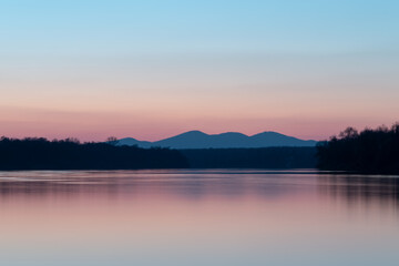Landscape with river, riparian forest silhouette and distant mountain under clear blue sky with pale red glow at horizon during twilight, pastel colors