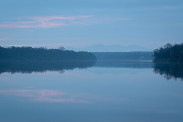 Fototapeta na wymiar Peaceful atmospheric landscape with pastel colors, Sava river at twilight, forested banks lead to distant mountain silhouette in haze, calm nature and water reflection