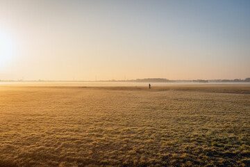 It is early in the morning in a Dutch polder. The grass is a little frosted and the morning mist hangs over the fields. The sun is just rising and in the distance a woman is running on a country road.