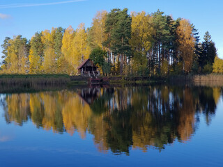 Fototapeta na wymiar small wooden house on the lake next to the autumn yellow forest. Reflection in the water of the lake.