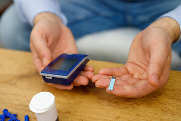 Man sitting at the table holding in his hands glucometer and stripes for checking blood sugar level, diabetes concept.