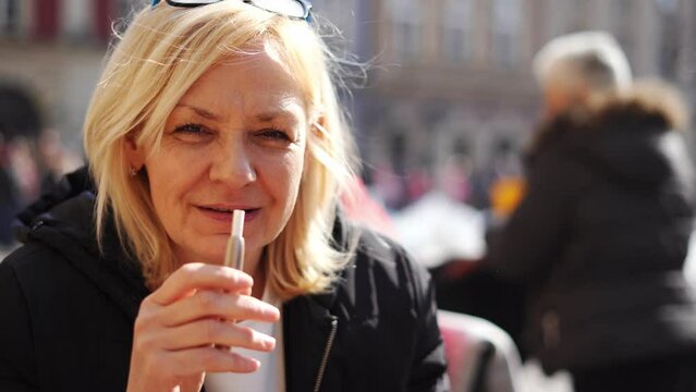 Middle aged blonde European woman smoking heated tabacco e-cigarette outdoors on the street. Alternative smoking way
