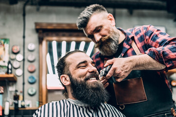 The barber trims the beard of the customer with shaving machine.