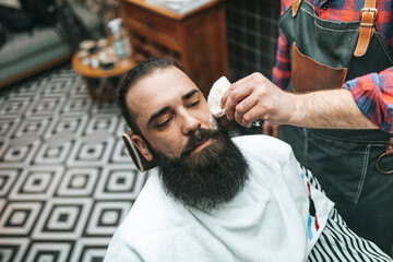 The middle-aged barber trims the beard of the customer in his barber shop. Trendy and stylish beard styling and cut.