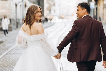 Naklejka premium An international wedding couple, a European bride and an Asian groom walk around the city together.
