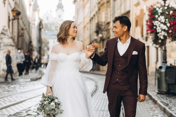 Naklejka premium An international wedding couple, a European bride and an Asian groom walk around the city together.
