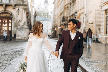 Naklejka premium An international wedding couple, a European bride and an Asian groom walk around the city together.