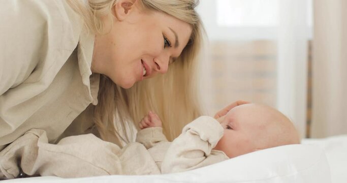 A cute baby lying on a white blanket, wearing white clothes. A young beautiful mother playing with baby, kissing, looking, smiling. The concept of caring for a newborn.