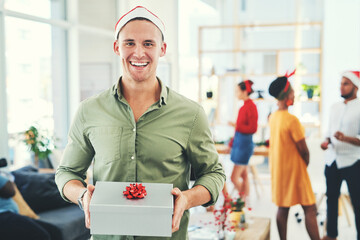Secret Santa is my favorite office tradition. Portrait of a handsome young businessman holding a gift box at an office Christmas party with colleagues in the background. - obrazy, fototapety, plakaty