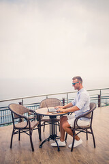 Handsome successful young male businessman sitting at a table by the pool with a laptop overlooking the Mediterranean Sea. Remote work on vacation. Vacation concept