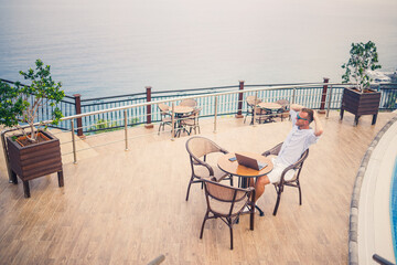 Handsome successful young male businessman sitting at a table by the pool with a laptop overlooking the Mediterranean Sea. Remote work on vacation. Vacation concept