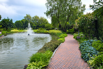 Fototapeta na wymiar A pond filled with water with a spray jet fountain in a park with pedestrian sidewalks made of stone tiles among different plants, landscape design of flower beds and trees.