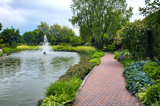 A pond filled with water with a spray jet fountain in a park with pedestrian sidewalks made of stone tiles among different plants, landscape design of flower beds and trees. - Powered by Adobe