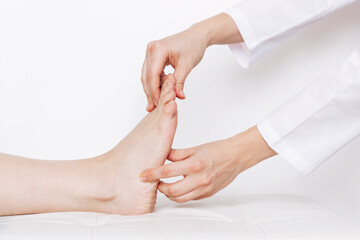 Examination of a young woman by an orthopedist. Cropped shot of female doctor holding a girl's foot in her hands on a white background. Flat feet, injury. Foot treatment. Pain from uncomfortable shoes - obrazy, fototapety, plakaty