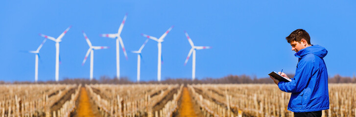 Banner. Young engineer takes notices on his notebook about wind turbines next to a vineyard.