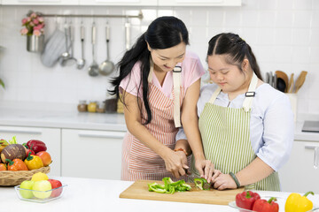 mother cutting fresh green peppers on wooden cutting board with down syndrome teenage girl or her daughter in the kitchen