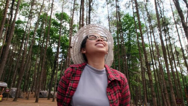 Hipster woman in straw hat and red plaid shirt is walking in a pine alley enjoying nature. Happy Asian female traveler sightseeing in pine tree garden.