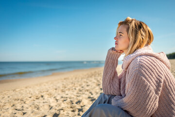 Fototapeta na wymiar Charming young woman in knitted sweater sitting on sandy beach at sunny day