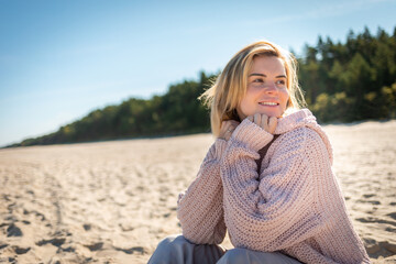 Charming young woman in knitted sweater sitting on sandy beach at sunny day