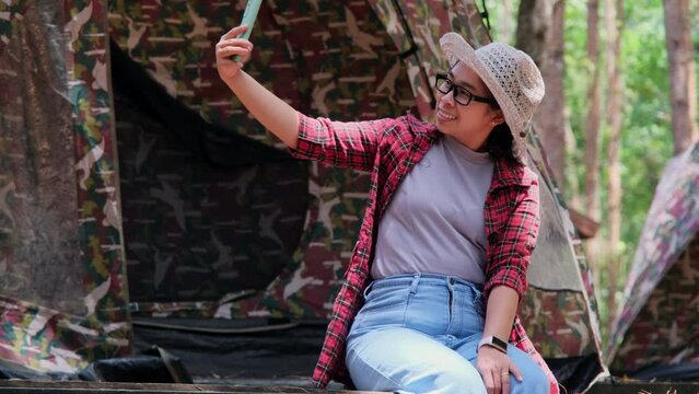 Beautiful Asian woman sitting under pine trees and taking selfie with smartphone in autumn forest on tent background. Camping in nature