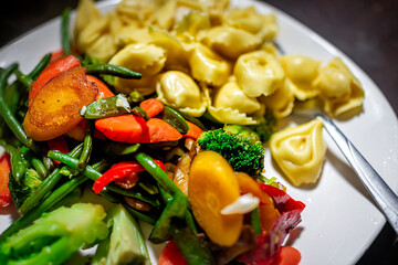 Dinner white plate vegetable dish with stiry-fry mix of carrots, broccoli red peper greens and sauce tortellini cheese filling pasta on table background macro closeup