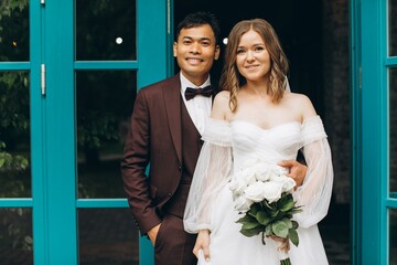 Fototapeta premium Wedding day. Beautiful European bride and her Asian groom posing against the backdrop of cafes and large windows.