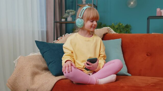 School kid at home alone dancing, moving to rythm, singing. Portrait of Caucasian funny little happy child girl sitting on sofa while listening music in headphones on smartphone player in living room.