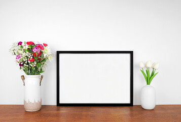 Mock up landscape black frame with vases of cut flowers. Wooden shelf against a white wall. Copy space.