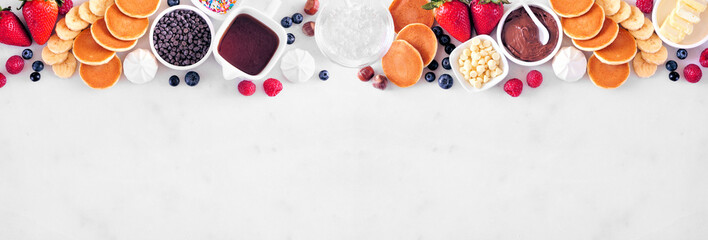 Breakfast or brunch pancake buffet top border. Overhead view on a white marble banner background. Mini pancakes with an assortment of toppings.